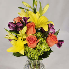 Bouquet of yellow lilies, orange roses, and purple flowers in a clear glass vase