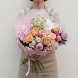 Handheld bouquet of pink, peach, and white flowers wrapped in pale pink paper