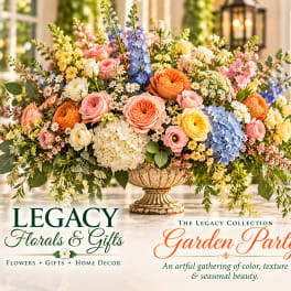 Large mixed floral arrangement in a decorative urn with pink, blue, yellow, and white blooms.