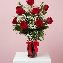 Red roses arranged in a glass vase with white filler flowers and a red ribbon