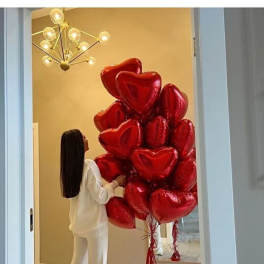 Cluster of red heart-shaped balloons in a room with a woman standing beside them.