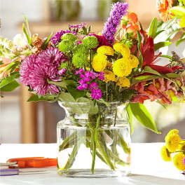 Colorful mixed bouquet of yellow, pink, green, and purple flowers in a clear glass vase.