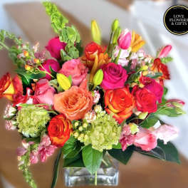 Vibrant arrangement of pink, orange, and yellow roses with lilies and green hydrangeas in a clear glass vase.
