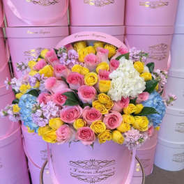 Pink and yellow roses arranged in a large pink hat box with white hydrangeas.
