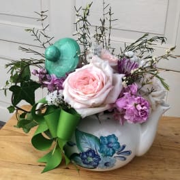 Pastel pink and purple flower arrangement in a painted ceramic teapot with green bow and tilted lid