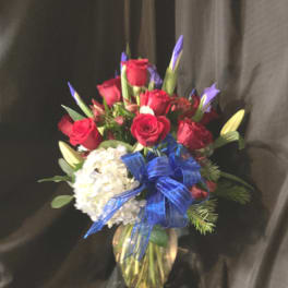 Red rose and purple iris arrangement with white hydrangea and blue bow in a glass vase