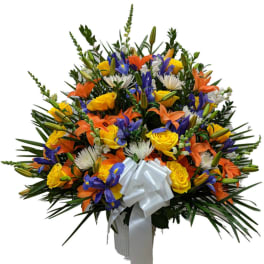 Tall fan-shaped arrangement of yellow roses, orange lilies, blue irises and white flowers with a white bow