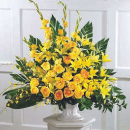 Tall yellow lily and rose arrangement with large green leaves in a white urn