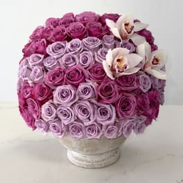 Round bouquet of pink and lavender roses with white orchids in a pedestal vase