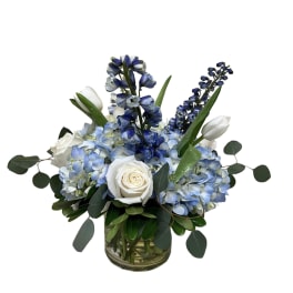 Blue hydrangeas, white roses, white tulips, and blue delphinium arranged in a clear glass cylinder vase