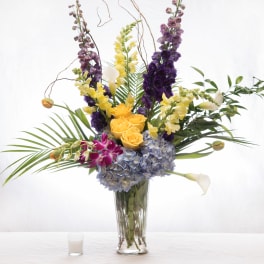 Tall mixed arrangement with blue hydrangeas, yellow roses, snapdragons, orchids, and calla lilies in a glass vase.