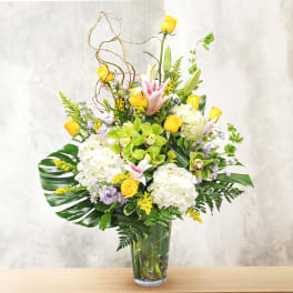 Tall mixed bouquet in a clear glass vase with yellow, white, pink, and green flowers.