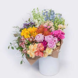 Large mixed-color bouquet with sunflowers, roses, peonies, and protea wrapped in kraft paper in a white cylinder.