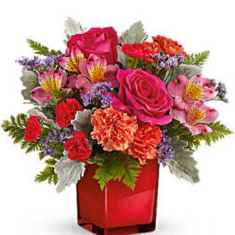 Compact arrangement of pink roses, orange carnations, and mixed blooms in a red cube vase