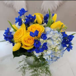 Low arrangement of yellow roses and tulips with blue delphinium and hydrangea in a clear glass vase