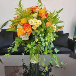 Tall vase of yellow and orange roses with white blooms and trailing ivy