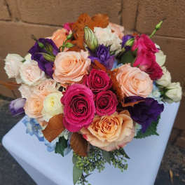 Round bouquet of pink and peach roses with purple lisianthus and blue accents on a white pedestal