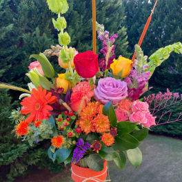 Tall mixed bouquet of bright roses, gerbera daisies, and mums in a glass vase wrapped with orange ribbon.
