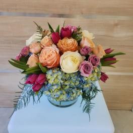 Short round arrangement of peach and cream roses, pink tulips, and blue hydrangeas in a clear glass vase
