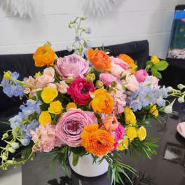 Bright mixed arrangement of pink, yellow, orange, and blue flowers in a white vase on a dark table