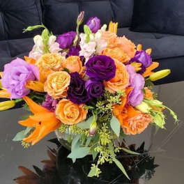 Low arrangement of orange lilies and roses with purple blooms in a clear glass bowl on a black table