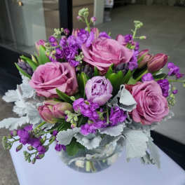 Compact arrangement of pink roses and purple flowers in a round glass vase