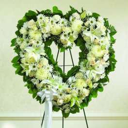 Heart-shaped standing wreath of white lilies, roses, and mums with green foliage and a white ribbon bow.