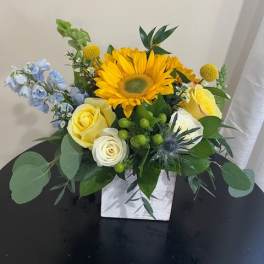 Bright sunflower and yellow roses with blue accents in a white cube vase