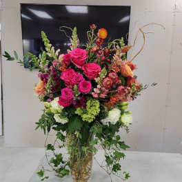 Tall arrangement of hot pink roses and mixed flowers in a clear glass vase with trailing ivy.