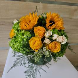 Round arrangement of yellow sunflowers and roses with green hydrangeas and cream spray roses.