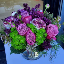 Compact arrangement of pink roses, purple blooms, and green hydrangeas in a clear glass vase
