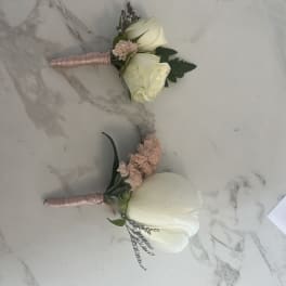 Two white rose boutonnieres with pink accents on a marble surface