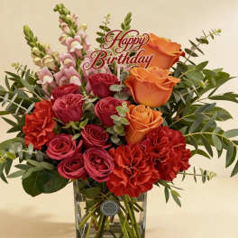 Birthday arrangement of red and orange roses, red carnations, and pink snapdragons in a glass vase.