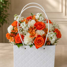 Orange and white flower arrangement in a white gift bag