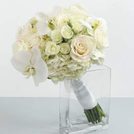 White bridal bouquet of roses, hydrangea, and orchids with ribbon-wrapped stems in a clear glass vase