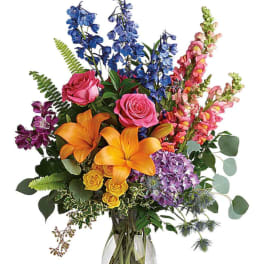 Colorful mixed bouquet in a clear glass vase with lilies, roses, and tall blue blooms
