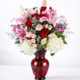 Bouquet of pink lilies, red roses, and white hydrangeas in a red glass vase