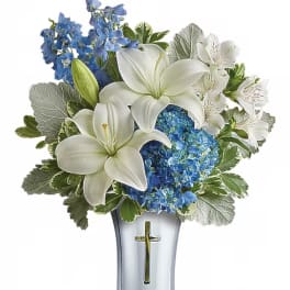 White lilies and blue flowers arranged in a clear vase with a cross