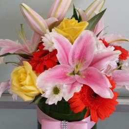 Pink lilies, yellow roses, and red gerbera daisies in a vase with a pink ribbon