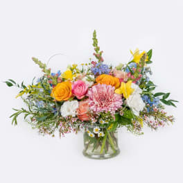Low glass vase of mixed pink, yellow, white, and blue flowers in a lush garden-style arrangement