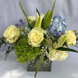 Arrangement of cream roses, lilies, and blue and green hydrangeas in a clear glass cube vase