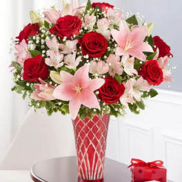 Red roses and pink lilies in a red glass vase beside a small gift box