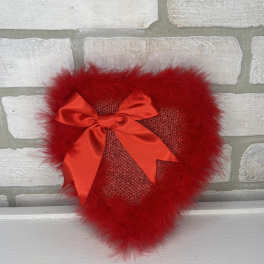 Red heart-shaped decorative pillow with feather trim and satin bow on a light brick background