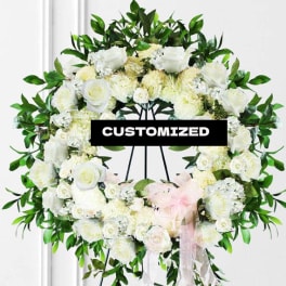 White floral wreath on a stand with green foliage and a pink ribbon
