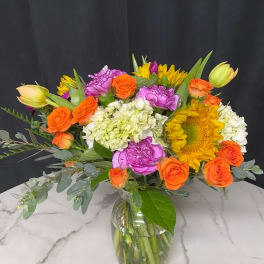Colorful bouquet of orange roses, sunflowers, and purple carnations in a glass vase