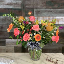 Mixed bouquet of bright pink roses, orange gerberas and yellow lilies in a clear vase with striped bow on a desk