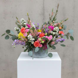 Low, wide arrangement of bright mixed flowers in a ribbed metal container on a white pedestal