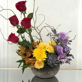 Mixed floral arrangement with red roses, yellow blooms, and purple flowers in a gray bowl vase