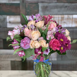 Mixed bouquet of peach roses and pink and purple blooms in a clear glass vase with blue ribbon