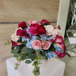 Low arrangement of pink roses, red ranunculus, blue hydrangeas, and dark pink calla lilies in a white vase